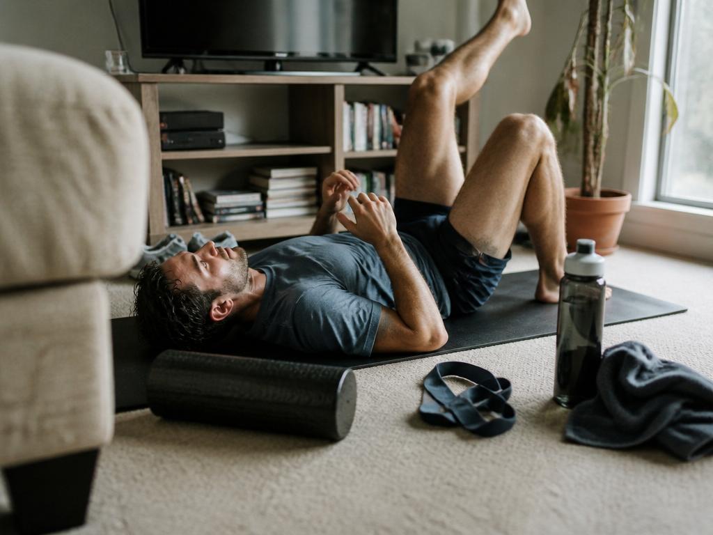 A man lies on his back on a thin yoga mat on the living-room floor doing a gentle leg-lift exercise, a water bottle and towel beside him.