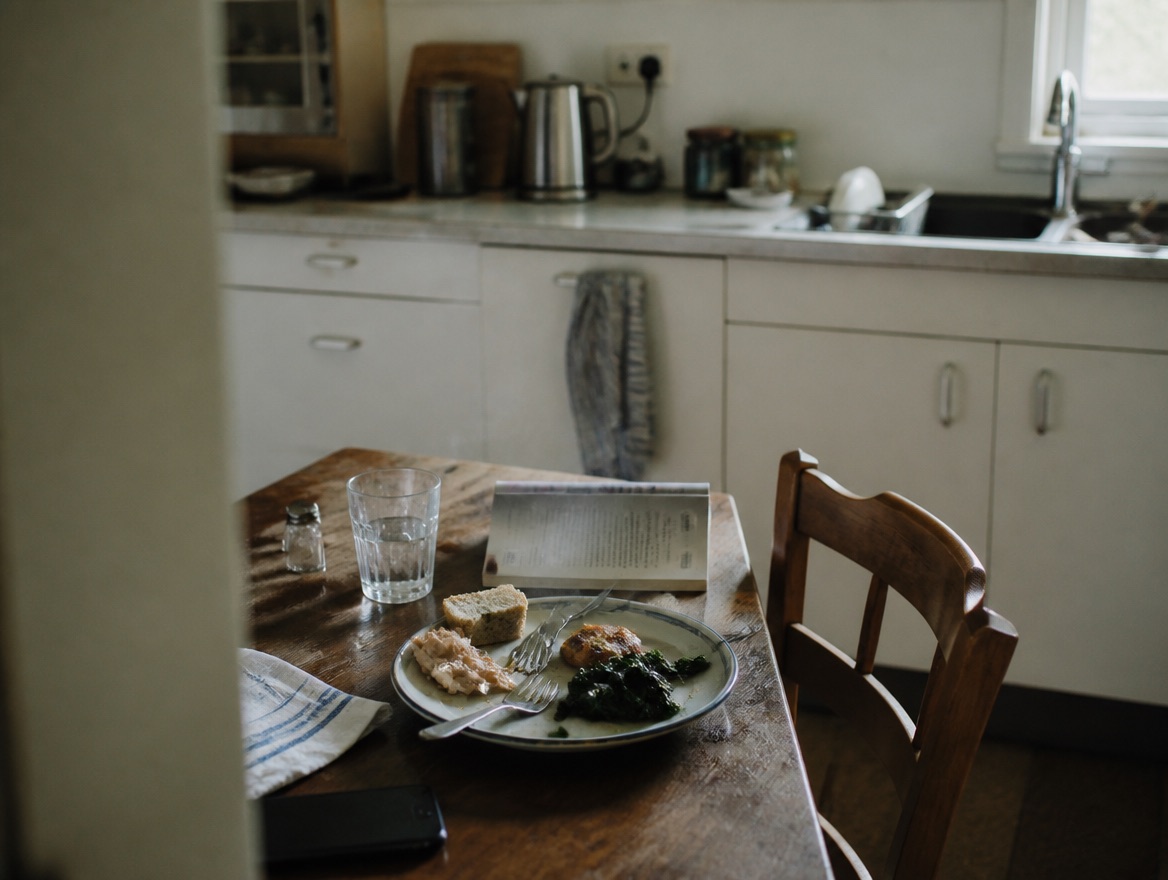A half-eaten plate of chicken, sautéed greens, and bread on a worn wooden kitchen table, with a glass of water, an open paperback face-down, a salt shaker, and a phone nearby; a wooden chair is pushed back at an angle and a lived-in kitchen with a kettle, sink, and blue-striped hanging tea towel sits soft-focus behind.