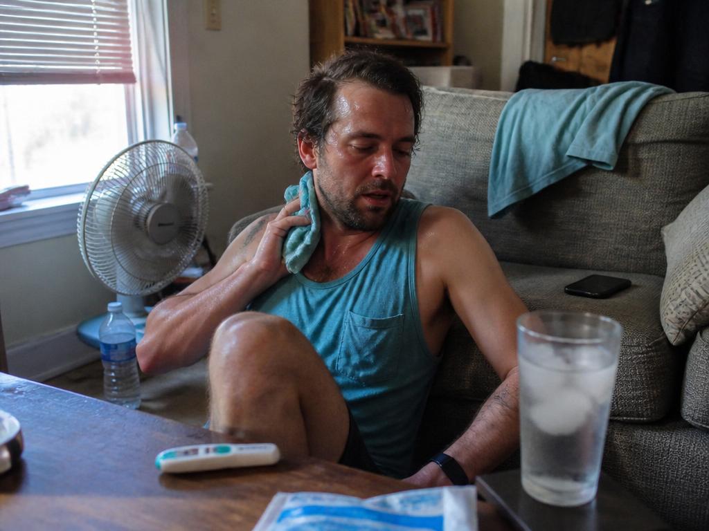 A man sits on the floor against a couch on a hot day, a small fan blowing on him, pressing a damp cooling cloth to his neck beside a glass of ice water.