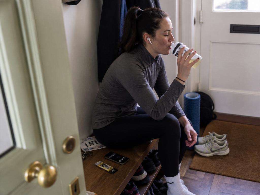 A woman in workout clothes sits on a bench by the front door drinking electrolyte mix from a sports bottle before a run, a torn sachet beside her.